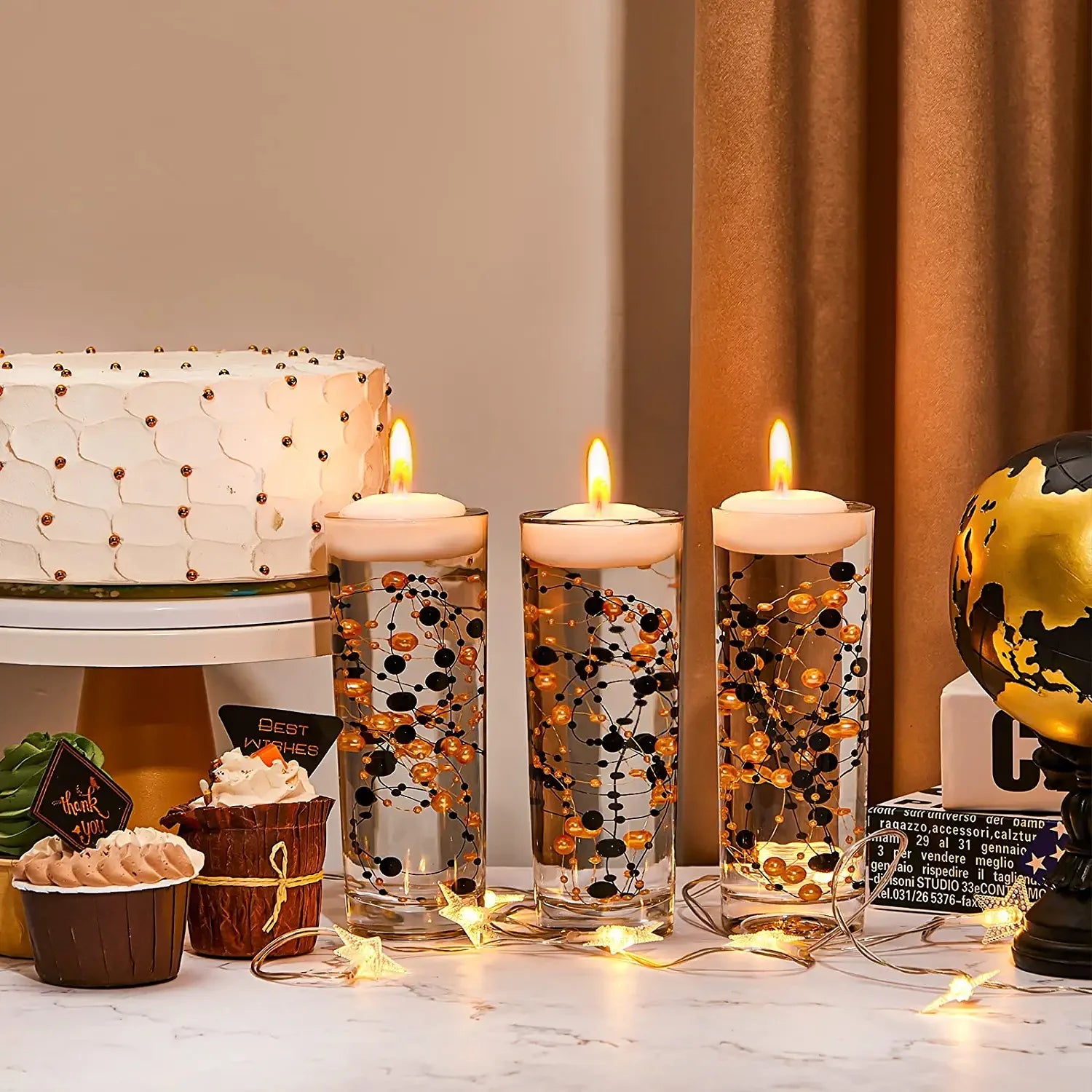 A table with a decorated cake, three lit candles in tall glasses, cupcakes, fairy lights, and a small globe. The candles are adorned with orange and black beads.