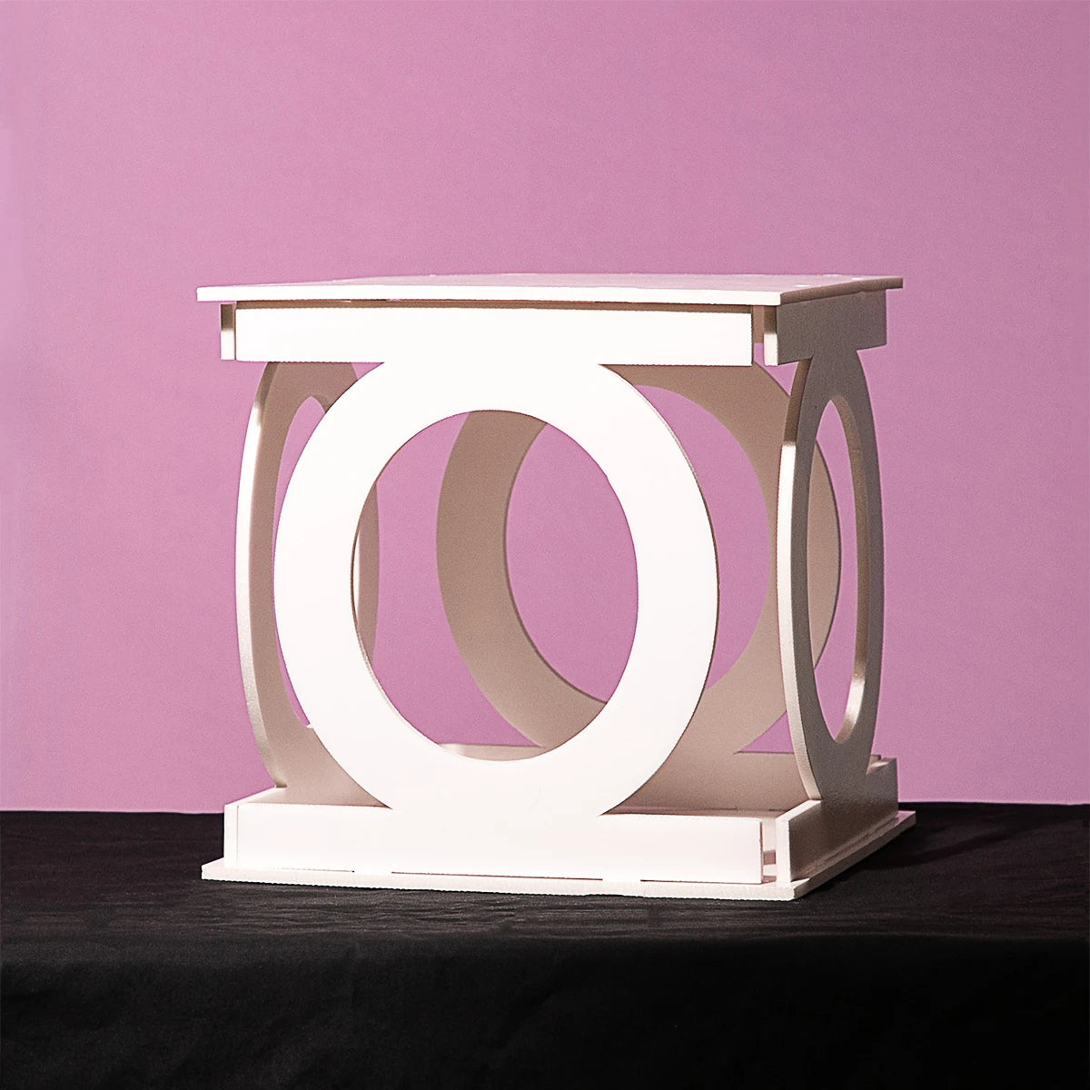 White geometric sculpture with circular cutouts, resting on a black surface against a pink background. The design features interlocking circles and square frames.