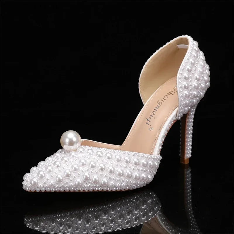 Elegant high heel shoe adorned with small white pearls and a larger pearl at the toe, set against a black background. The shoe features an open back and closed pointed toe.