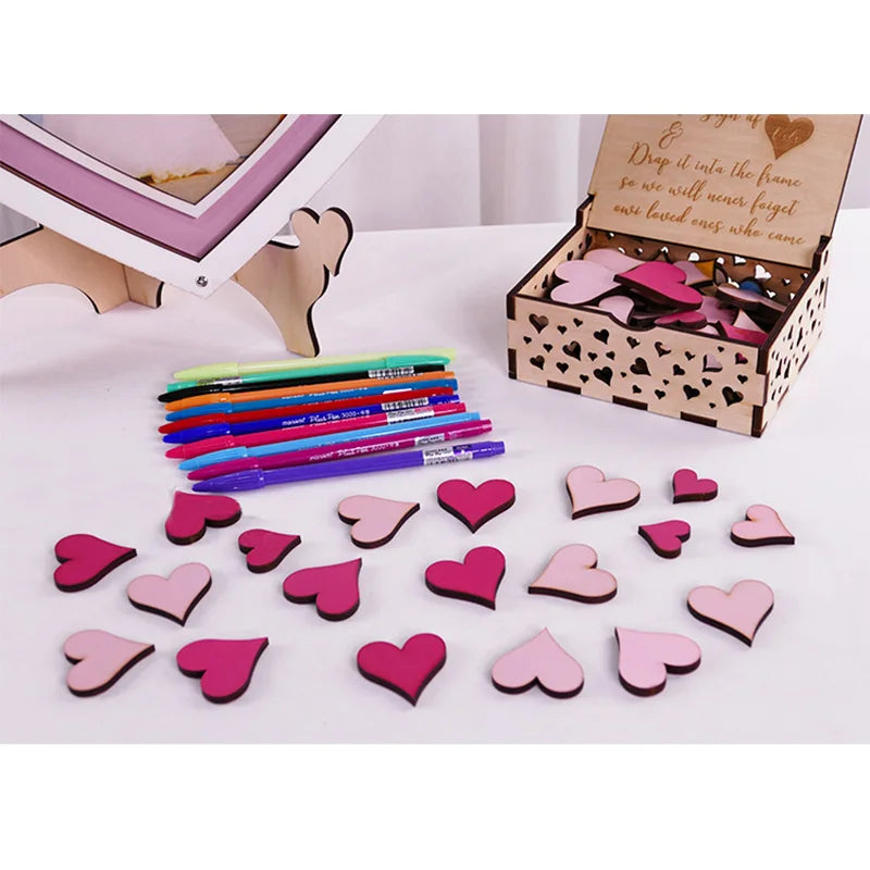 Box with pink and red heart shapes scattered on a table, alongside colorful pens. A wooden frame is in the background, and the box features heart cutouts.