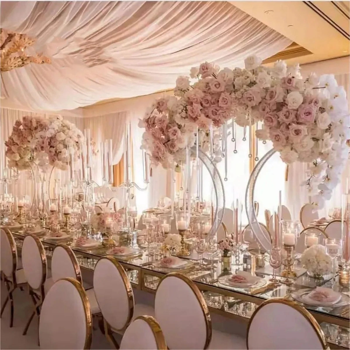 Elegant wedding reception with long tables, adorned with white and blush rose centerpieces, tall candles, and gold-accented chairs. Draped fabric ceiling enhances the romantic ambiance.