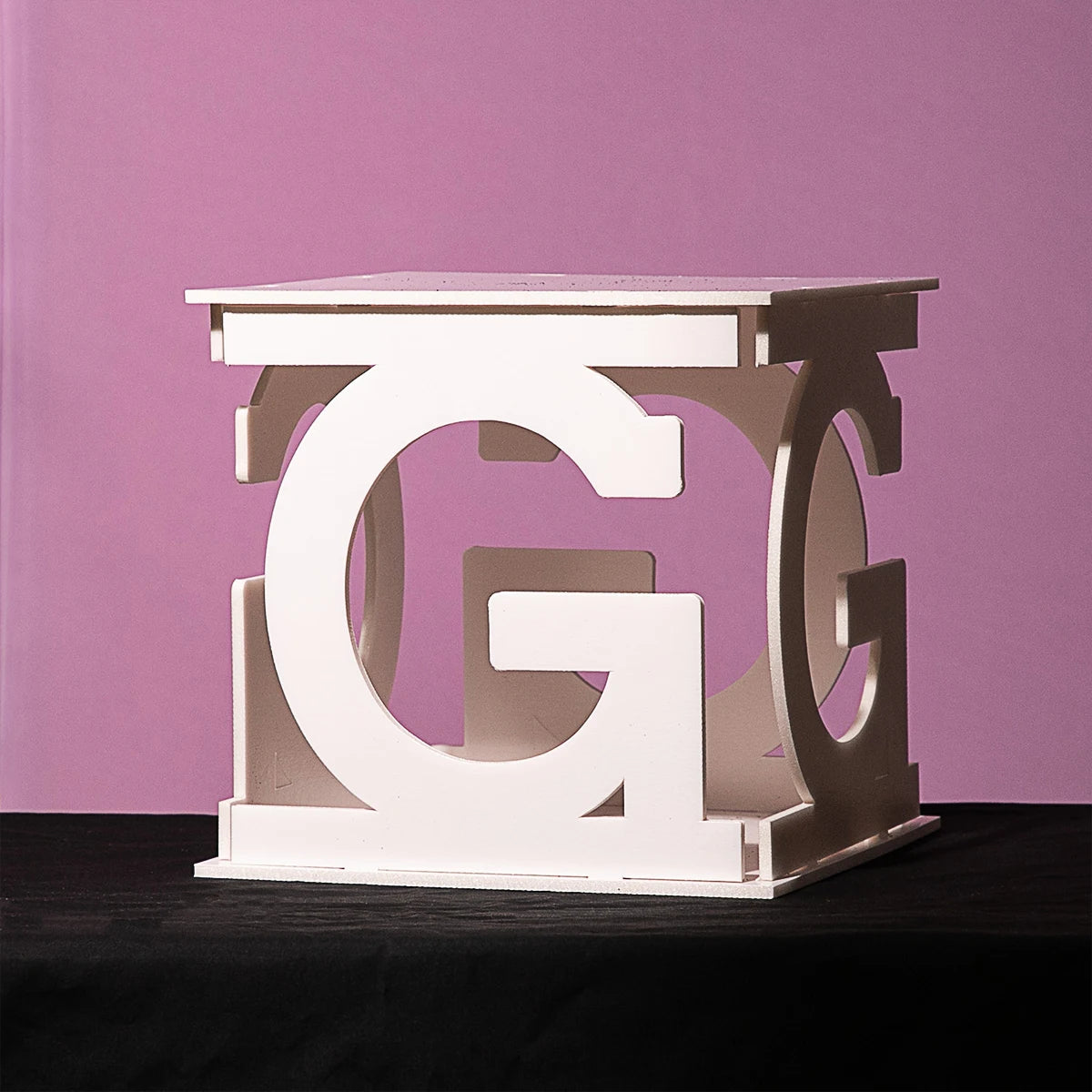 White cubic structure made of large, interlocking letter Gs against a pink background, resting on a black surface.