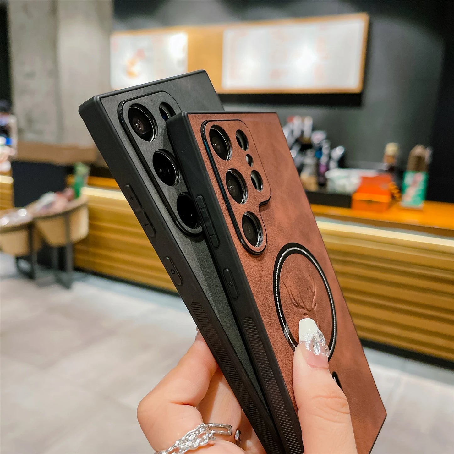 Two smartphones with brown and black protective cases, featuring multiple camera lenses, are held in a hand with a silver ring. A blurred cafe setting is in the background.