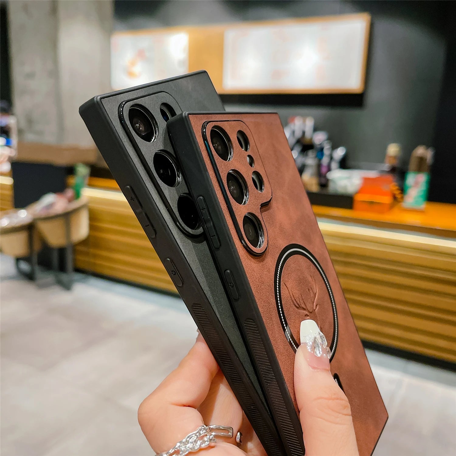 Two smartphones with brown and black protective cases, featuring multiple camera lenses, are held in a hand with a silver ring. A blurred cafe setting is in the background.