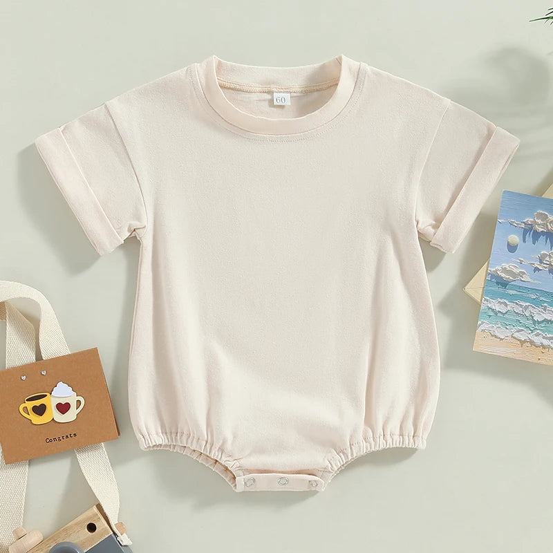 Cream-colored baby romper with short sleeves and snap buttons at the bottom, displayed on a flat surface. Nearby, a card with two smiling cups and a seaside postcard.