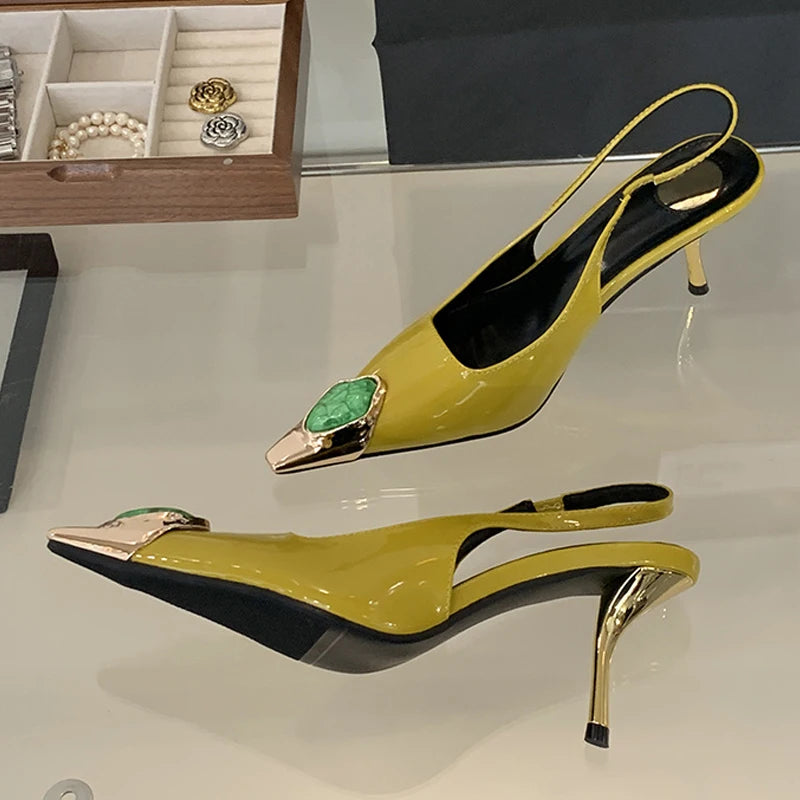 Yellow slingback high heels with gold pointed toe and green gem adornment displayed on a glass surface. Jewelry box with rings, bracelet, and pins in the background.