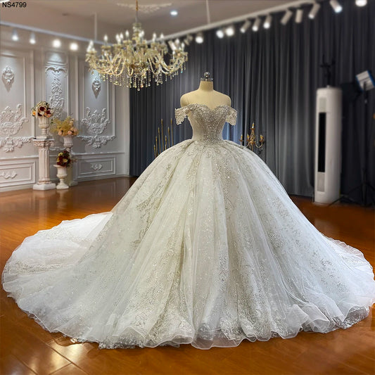 Elegant off-the-shoulder wedding gown on a mannequin, featuring intricate lace and beading. Set in a luxurious room with a chandelier and floral arrangements.