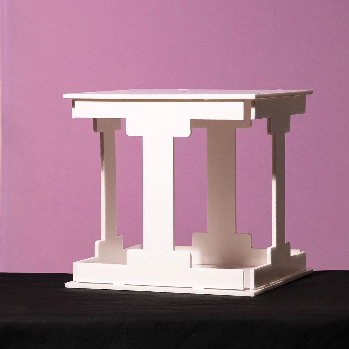 White geometric table with pillar-like legs against a purple background, resting on a dark surface.