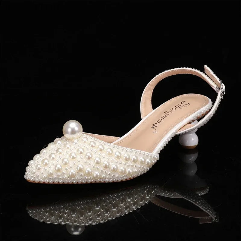 Elegant pearl-embellished slingback sandal with a pointed toe and chunky heel, featuring a prominent pearl accent at the toe, set against a black background.