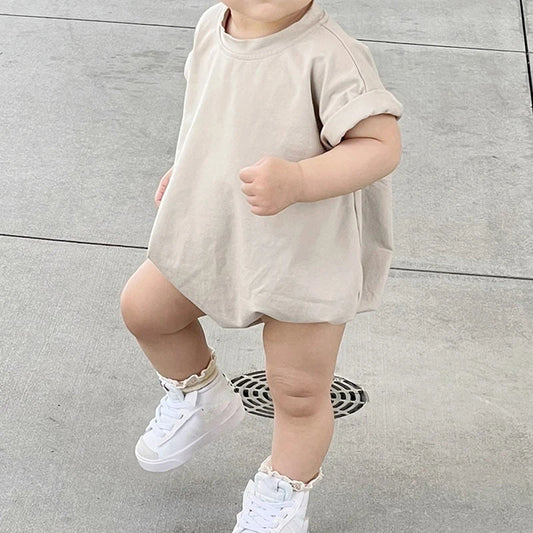 A toddler walks on a tiled surface, wearing a loose beige dress and white sneakers with frilly socks.