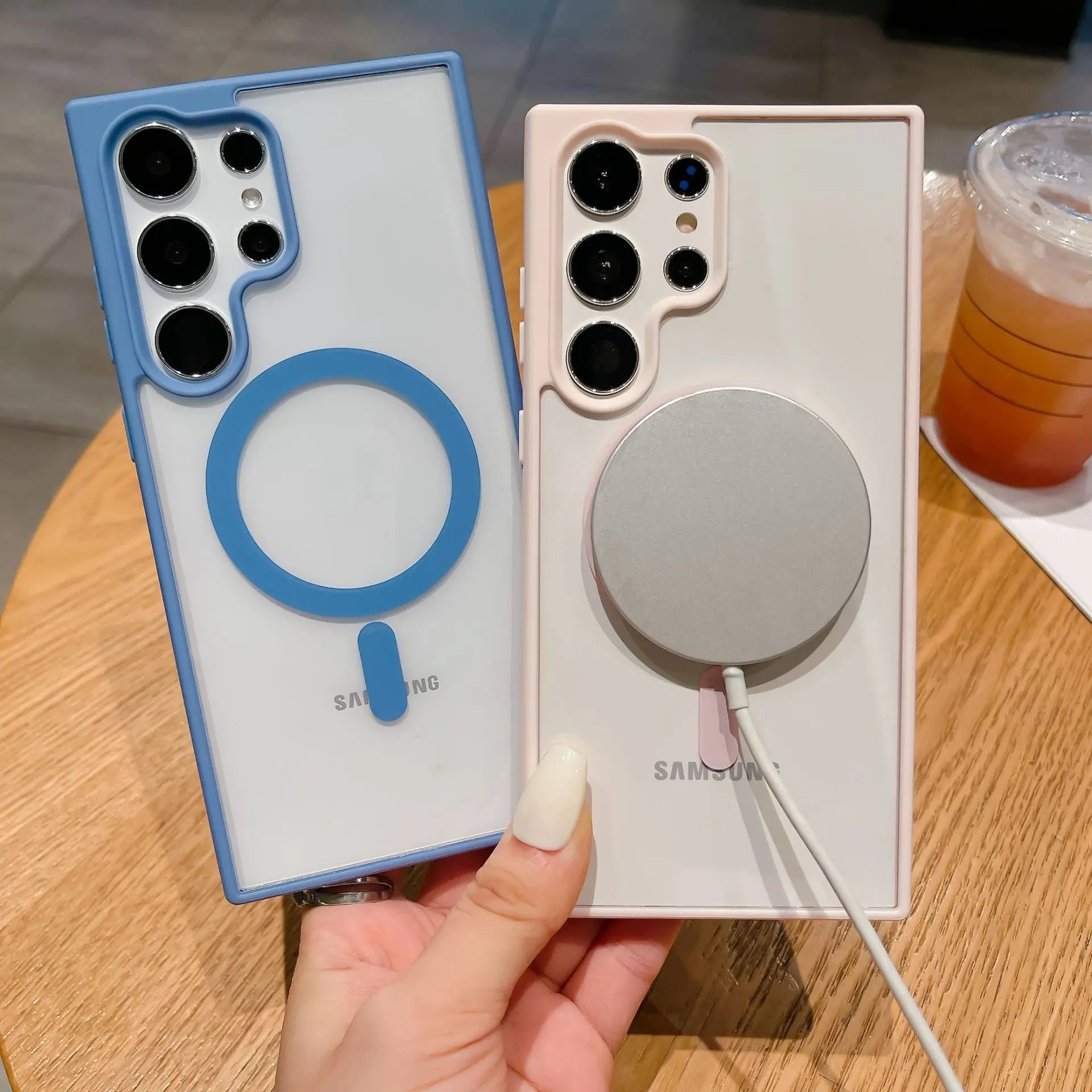 Two Samsung phones with protective cases are displayed. The left case is blue with a circular ring; the right case is white with a magnetic charger attached. A drink is in the background.