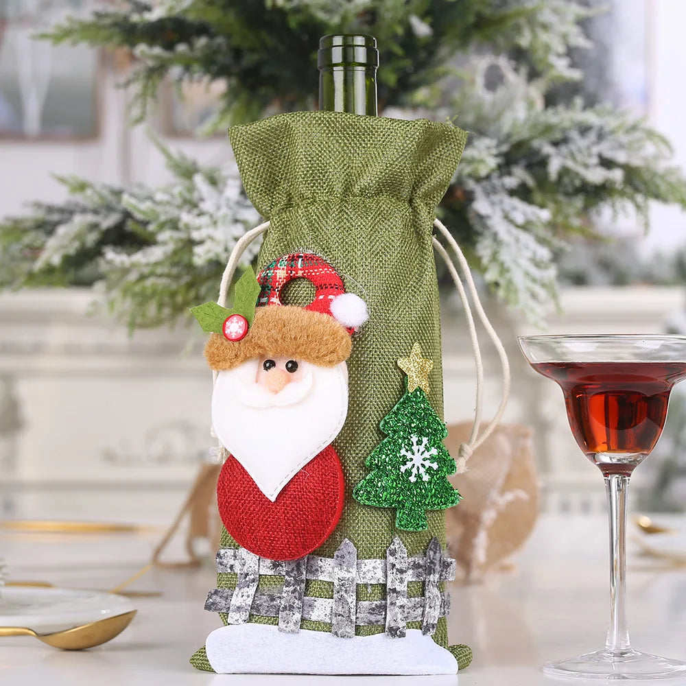 Green wine bottle cover with Santa and tree decorations beside a glass of red wine, set against a backdrop of a snow-dusted Christmas tree.