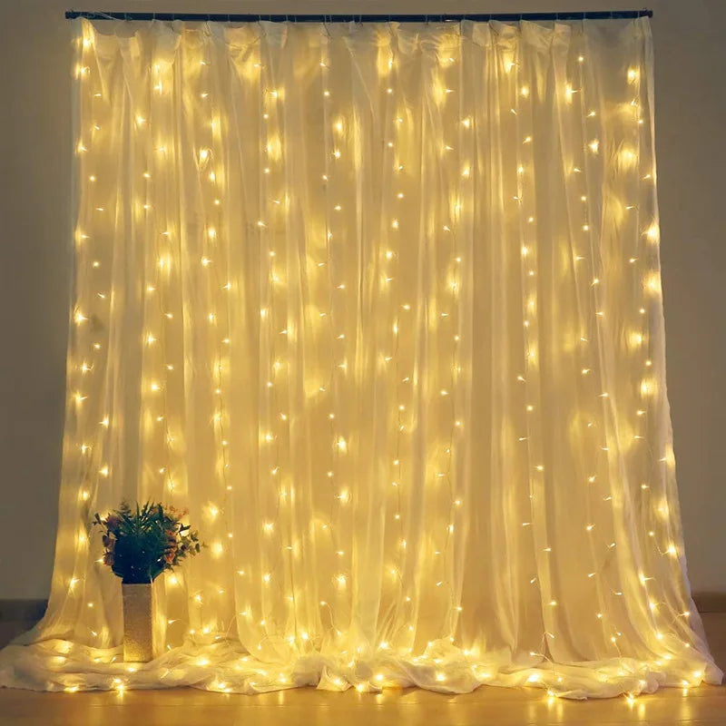 A curtain of warm yellow string lights hanging from a rod, creating a soft glow on sheer fabric; a pot of flowers sits at the base on a wooden floor.
