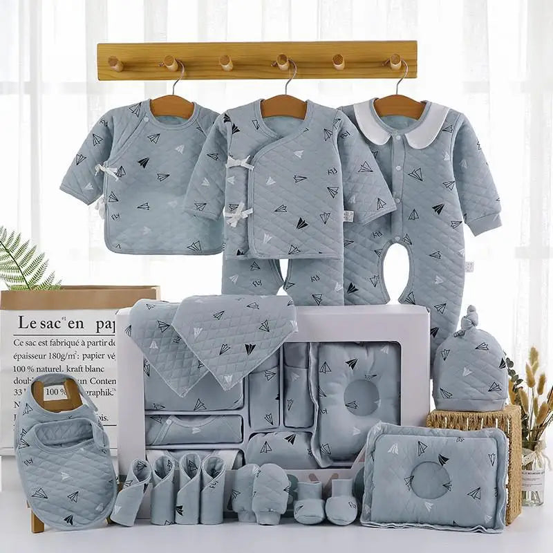 A set of baby clothes in gray with a paper airplane pattern. Includes outfits, bibs, a hat, mittens, booties, and a quilted pillow, neatly arranged on hooks and a display box.