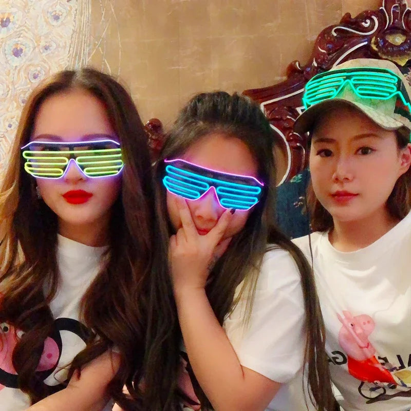 Three women wearing colorful LED shutter shades. Two make playful poses while sitting indoors in casual outfits with graphic prints, against a decorative background.