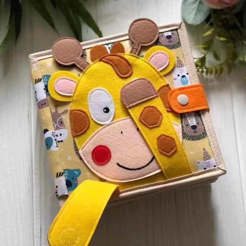 A cute fabric children's book with a giraffe face made of felt on the cover. The giraffe has a playful expression, with yellow spots and a foldable strap secured by a button.