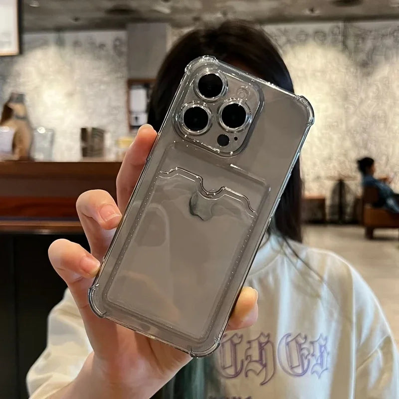 A person holds a smartphone with a clear case featuring a built-in card holder, showing the triple camera setup. They are in a room with blurred decor and another person in the background.
