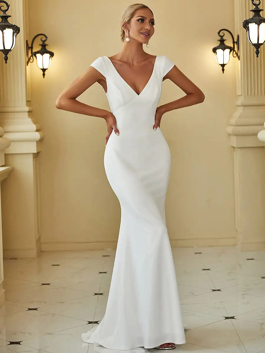 Woman in an elegant, form-fitting white gown with a deep V-neck, standing in a classical interior with soft lighting and ornate wall sconces, looking poised and graceful.