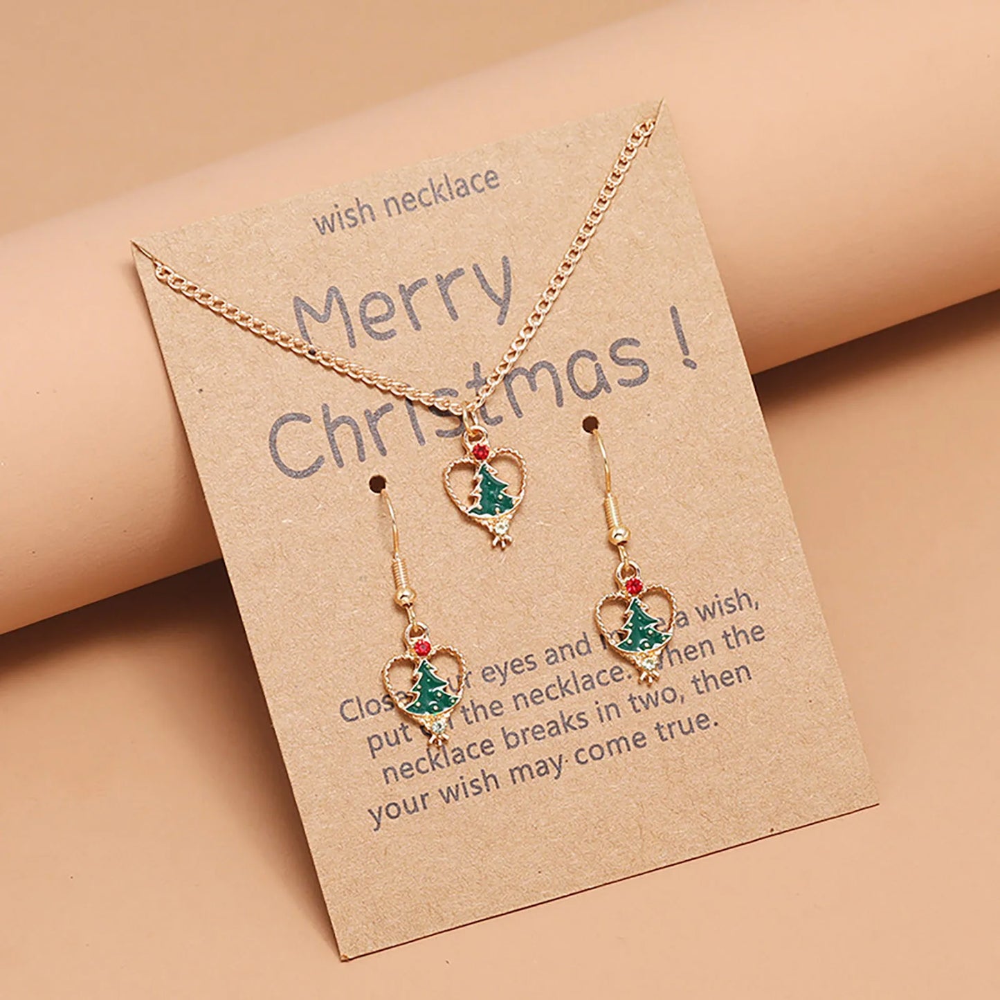 Gold necklace and earrings set with green Christmas tree and red gem accents on a card reading "Merry Christmas." Brown background and festive design.
