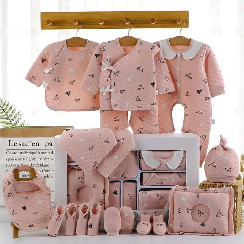 Set of pink baby clothes with airplane patterns, including rompers, bib, hat, gloves, and blanket, neatly arranged with a light background and a wooden rack above.