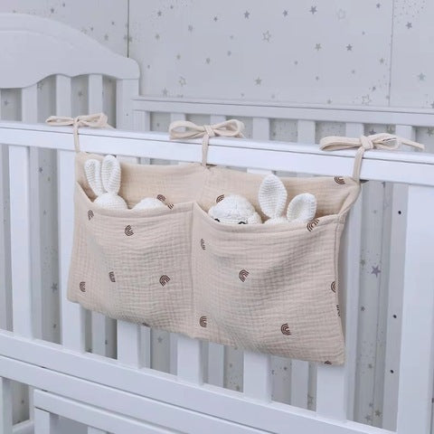 Fabric organizer with two pockets hangs on a white crib. It holds plush white bunny toys and has a subtle pattern. The background displays a star-themed wall.