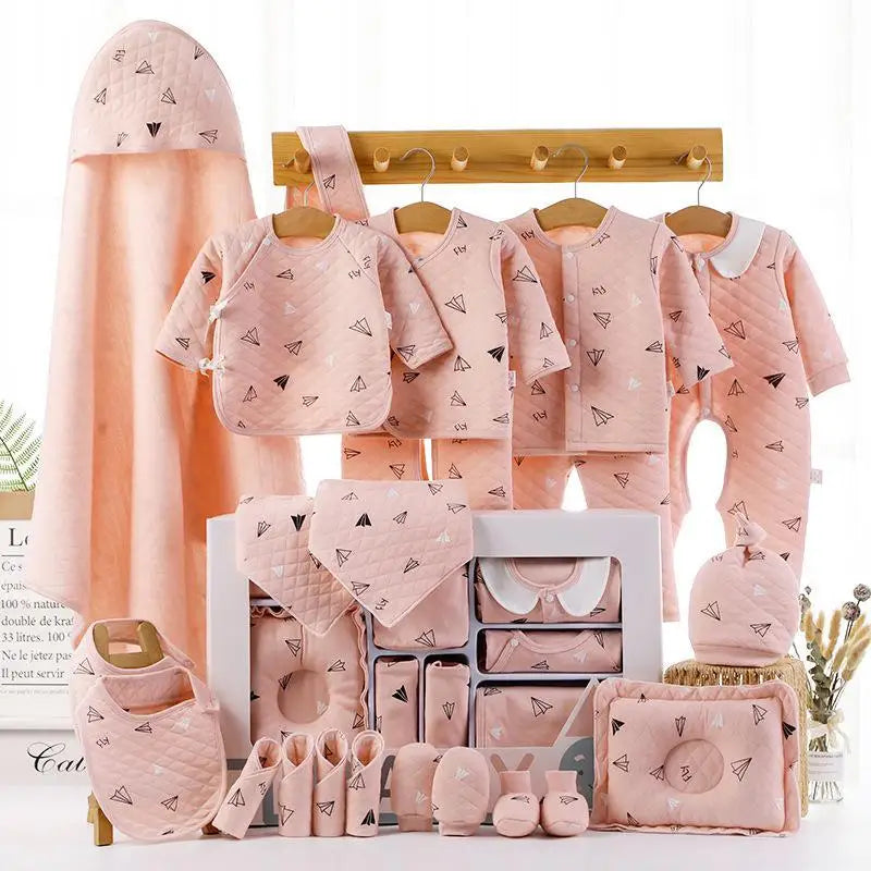 Set of pink baby clothes with paper plane patterns, including outfits, bibs, booties, and a hooded towel, displayed on a wooden rack against a light background.