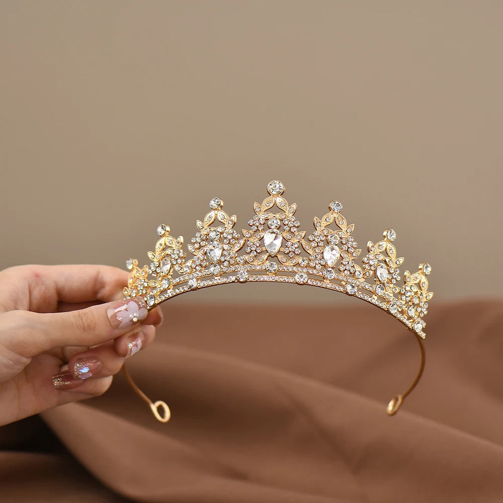 A hand holds an ornate gold tiara adorned with sparkling crystals and intricate detailing. The background is a soft brown fabric, enhancing the tiara's elegance.