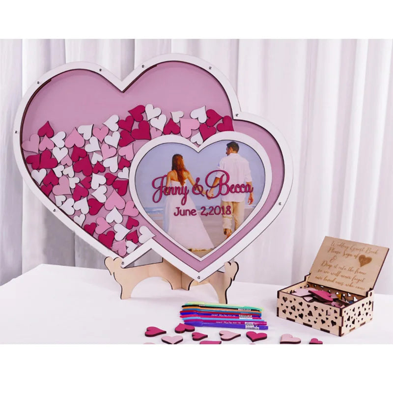 Wedding guest book display with two heart frames filled with pink and white wooden hearts. A couple's photo and "Jenny & Becca, June 2, 2018" are in the center. Pens and a heart box in front.