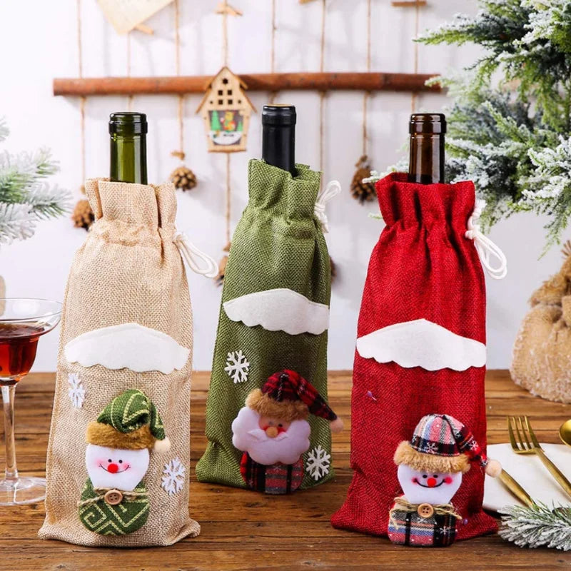 Three festive wine bottle covers in beige, green, and red, adorned with snowman and Santa motifs. They are set on a wooden table with Christmas decorations in the background.