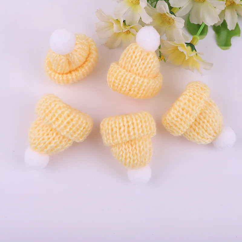 Five small, knitted yellow hats with white pom-poms are arranged on a white surface next to delicate, light yellow flowers.
