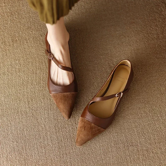 A pair of brown, pointed-toe flats with a buckle strap, one worn and one placed on a textured beige carpet. The wearer is wearing a green skirt.