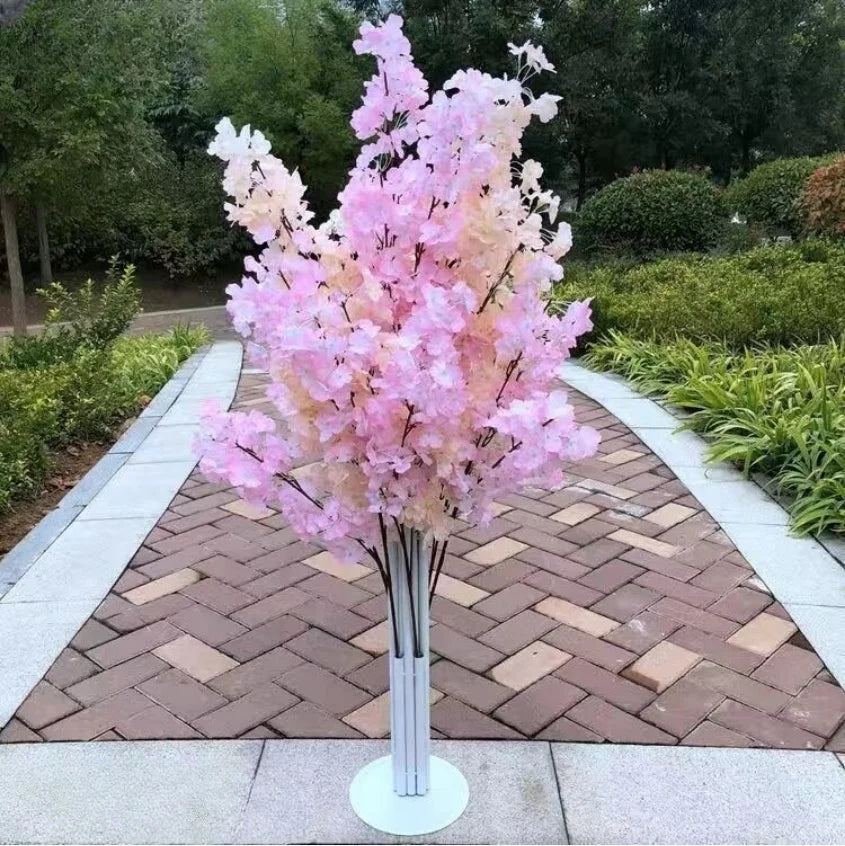 Artificial cherry blossom tree with pink and cream flowers on a brick pathway. Surrounded by greenery, creating a serene, garden-like environment.