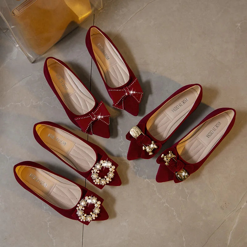 Three pairs of elegant red velvet flats with ornate embellishments: bows with pearls, crystals, and metallic charms, displayed on a light stone surface.