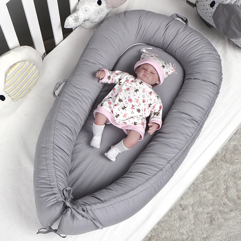 Baby sleeps in a gray infant lounger, wearing a floral outfit and pink hat. The scene is cozy, with stuffed animals nearby in a crib on a soft, textured rug.