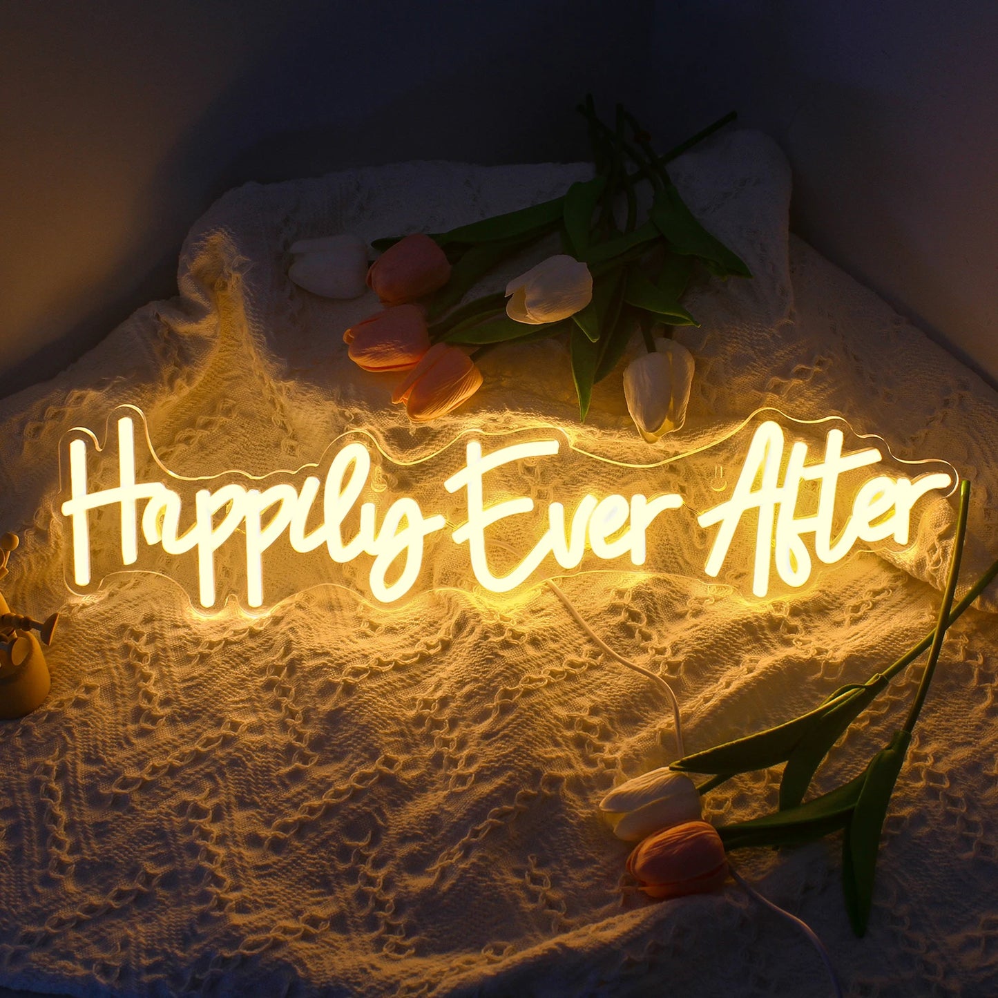 Glowing neon sign reading "Happily Ever After" on a textured fabric backdrop, surrounded by pink and white tulips.