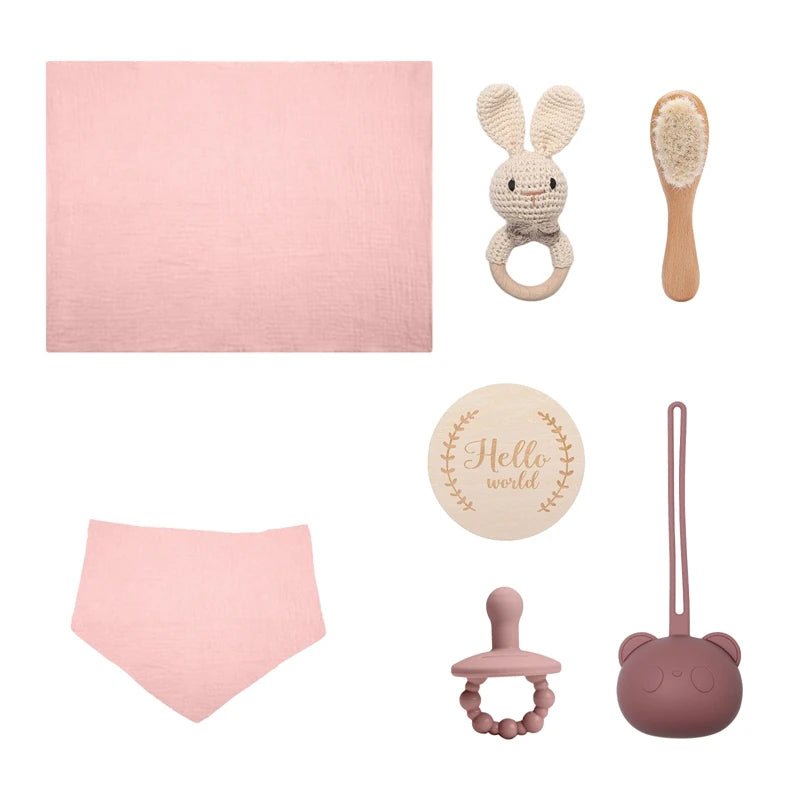 Pink and beige baby essentials set: blanket, bib, teething ring, hairbrush, pacifier, "Hello world" wooden plaque, and a bear-shaped storage case, all in soft colors.