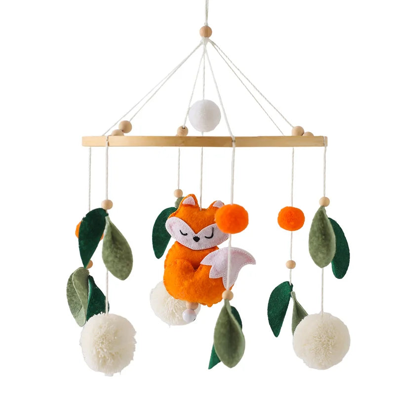 A baby mobile with a plush orange fox, white pom-poms, and green felt leaves, hanging from a wooden frame with strings.
