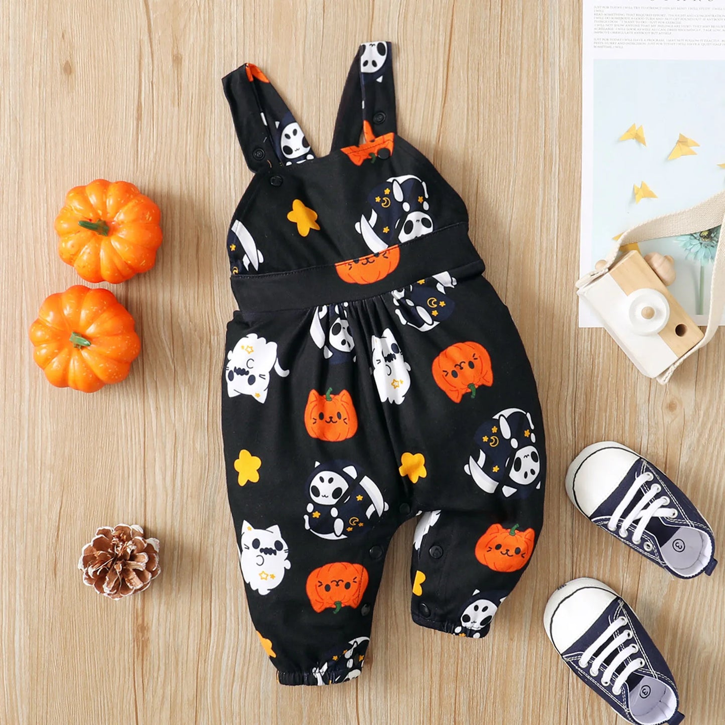 Black toddler overalls with Halloween-themed prints of pumpkins, ghosts, and cats. Surrounding items include mini pumpkins, a wooden camera, and sneakers on a wooden surface.