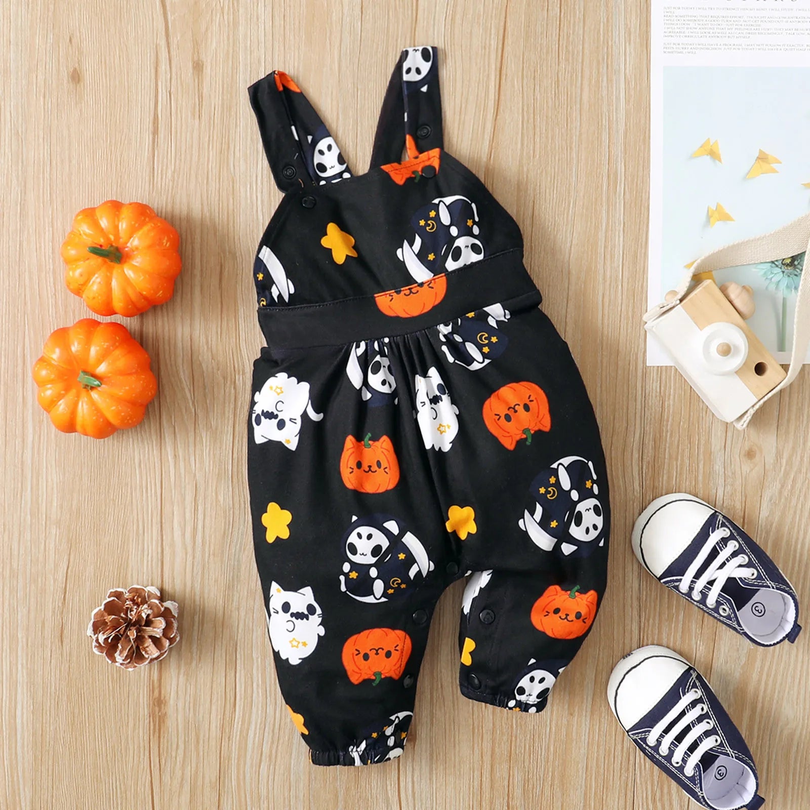 Black toddler overalls with Halloween-themed prints of pumpkins, ghosts, and cats. Surrounding items include mini pumpkins, a wooden camera, and sneakers on a wooden surface.