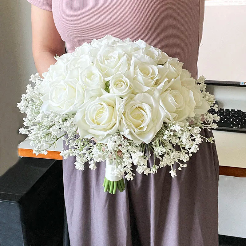 Woman in a pink top holds a bouquet of white roses and baby's breath, standing indoors near a desk with a keyboard partially visible.
