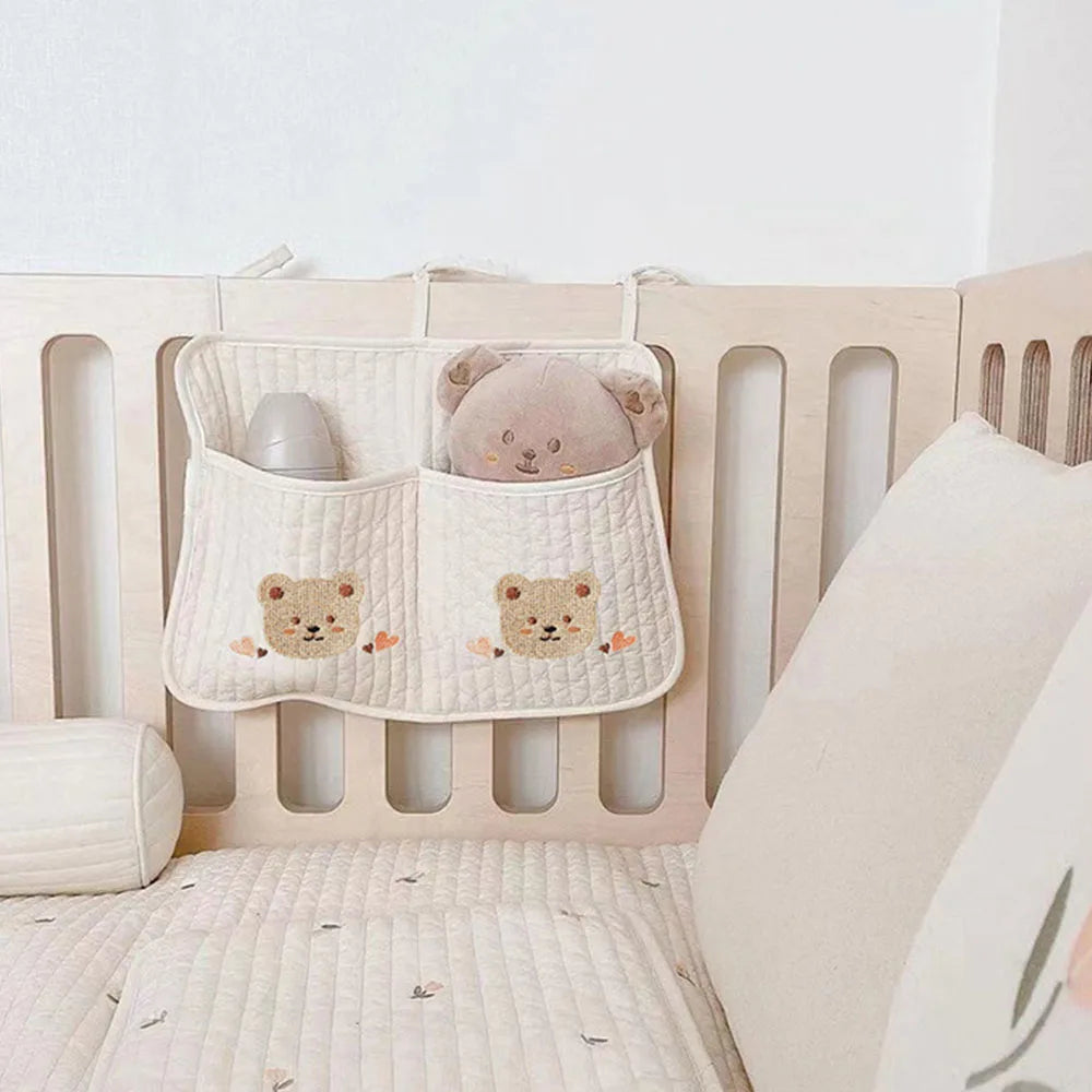 A wooden crib features a quilted organizer with two pockets, decorated with bear faces. It holds a soft toy bear and a grey bottle, next to a cream quilt and pillow.