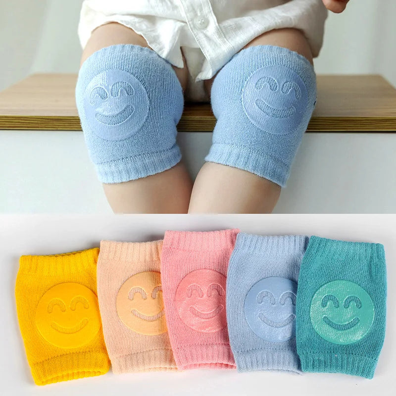 Baby wearing blue knee pads with smiley faces, sitting on a wooden surface. Below, five colorful knee pads in yellow, peach, pink, blue, and teal, each with a smiley face design.