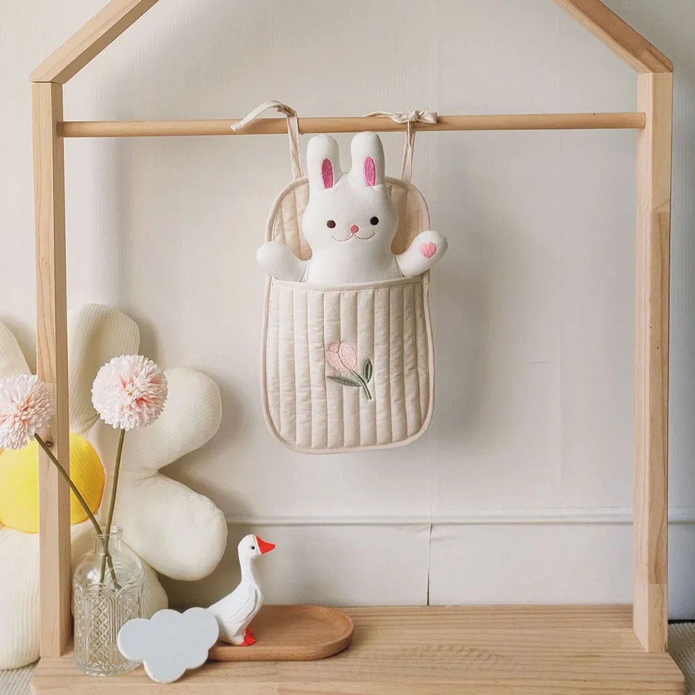 A soft white bunny toy with pink ears sits in a hanging beige quilted pouch with floral embroidery. Nearby are white and pink flowers and a ceramic duck on a wooden stand.