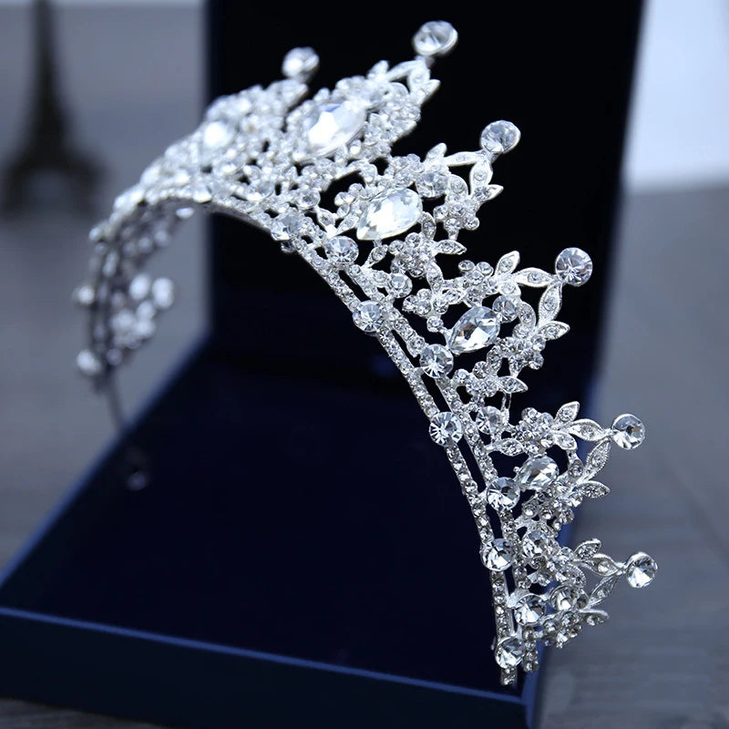 Elegant silver tiara adorned with sparkling rhinestones and intricate floral patterns, displayed on a dark blue cushion.