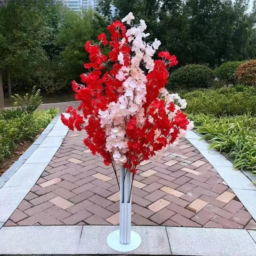 A decorative tree with vibrant red and pale pink blossoms, in a garden setting with paved walkway, surrounded by green bushes and tall trees in the background.