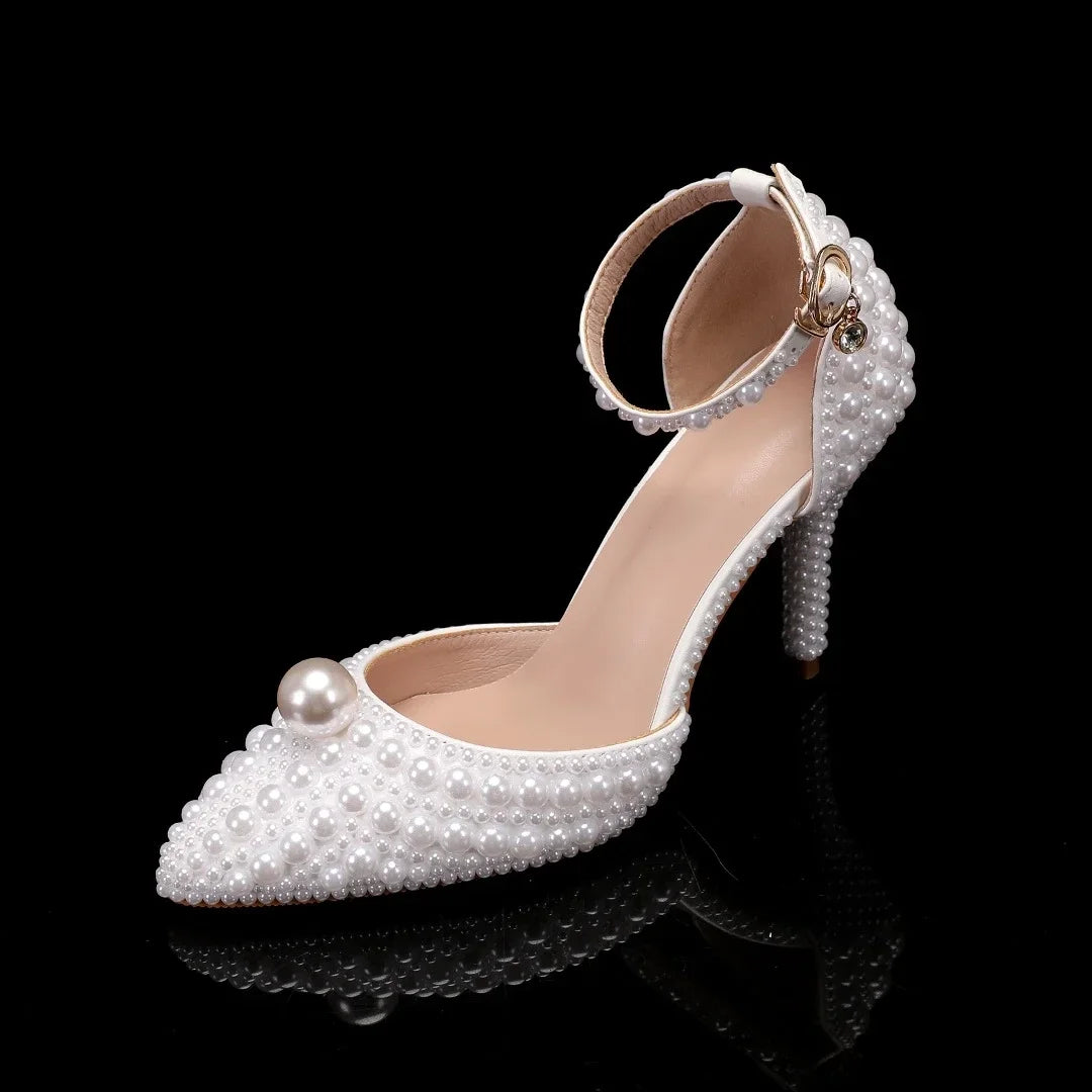 Elegant white high heel with ankle strap, adorned with pearls and a large pearl on the toe, displayed on a black reflective surface.