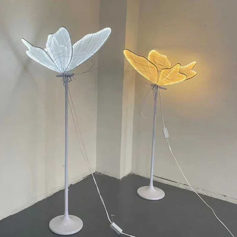 Two standing lamps with leaf-shaped shades; one emits cool white light, the other warm yellow, both on white bases against a plain wall in a minimalist setting.