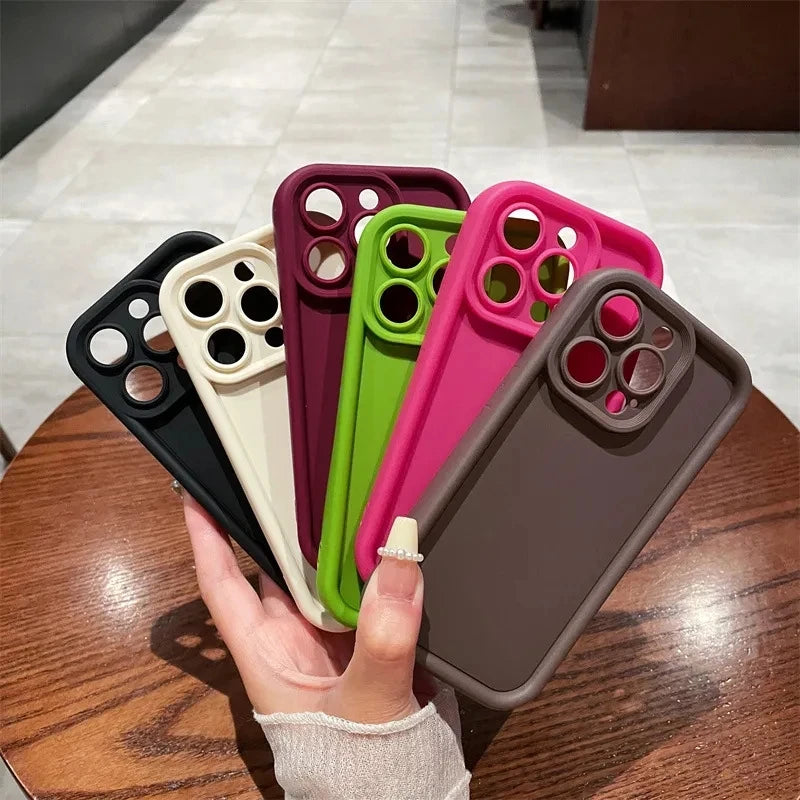 Hand holding six colorful smartphone cases, fanned out on a wooden table. The cases are black, cream, maroon, green, pink, and brown, each with camera cutouts.