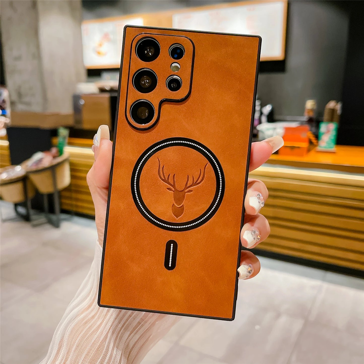 A tan leather-backed phone case features multiple camera lenses and a circular deer emblem. The device is held in a hand with long, manicured nails in a modern cafe setting.