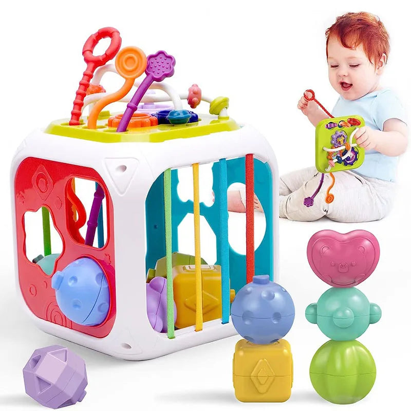 A baby plays with a colorful activity cube featuring beads, shapes, and a maze. Beside it are stacking shapes in pink, blue, yellow, and green.